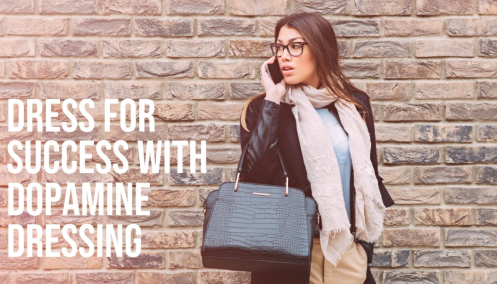 A person standing against a brick wall holding a black handbag, with their face obscured by a pixelation effect. The person is wearing a beige skirt, black jacket, and white scarf.