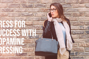 A person standing against a brick wall holding a black handbag, with their face obscured by a pixelation effect. The person is wearing a beige skirt, black jacket, and white scarf.