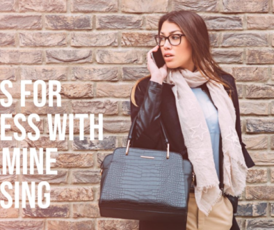 A person standing against a brick wall holding a black handbag, with their face obscured by a pixelation effect. The person is wearing a beige skirt, black jacket, and white scarf.