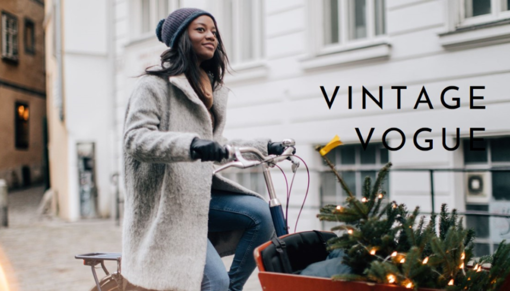 A person with their face obscured is riding a bicycle with a basket containing greenery and what appears to be a small Christmas tree, on a cobblestone street beside white buildings.