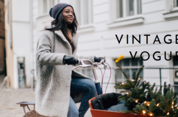 A person with their face obscured is riding a bicycle with a basket containing greenery and what appears to be a small Christmas tree, on a cobblestone street beside white buildings.